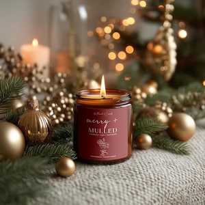 May include: A lit candle in a glass jar with a red label that reads "merry + MULLED". The candle is surrounded by gold ornaments, pine needles, and a soft, neutral-colored blanket. The background features blurred lights, suggesting a festive holiday theme.