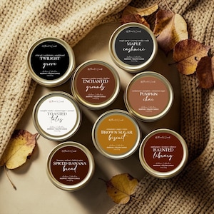 May include: A collection of eight round beeswax and tallow candles in gold metal tins, each featuring a unique label with scent notes and names like Twilight Grove, Maple Cashmere, Enchanted Grounds, Pumpkin Chai, Toasted Tales, Brown Sugar Biscuit, Spiced Banana Bread, and Haunted Library, each weighing 57 g.