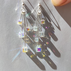 Op de afbeelding: Een paar zilveren oorhangers met iriserende kubusvormige kristallen die aan meerdere strengen hangen.