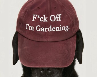 May include: A maroon baseball cap featuring the white embroidered text F*ck Off I'm Gardening. The cap has a structured crown, a curved brim, and a button on top. It is positioned on the head of a black dog with visible ears and a snout.
