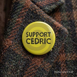 May include: A round yellow pinback button featuring a dark gray spiral pattern and the black text SUPPORT CEDRIC. The button has a diameter of 2.25 inches and is attached to a textured brown and black tweed fabric with a visible weave pattern.