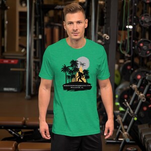 Puede incluir: Una camiseta verde con una silueta de Bigfoot caminando frente a una luna llena y palmeras. El texto "BIRDDOGDRONESERVICES.COM WILLISTON, FL" está impreso debajo del gráfico. La camiseta está hecha de un material suave.