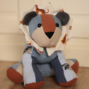 Può includere: Un leone di peluche realizzato con vari ritagli di tessuto nelle tonalità del blu, marrone e crema. Il leone presenta una criniera color crema con un motivo di piccoli leopardi arancioni e marroni, orecchie grigio scuro e naso nero con occhi a bottone.