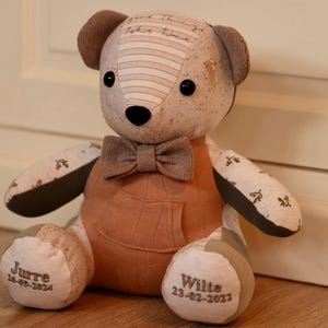 Puede incluir: Un oso de peluche de patchwork marrón y beige con una pajarita marrón. El oso tiene una nariz marrón y ojos negros. Los pies del oso están bordados con los nombres "Jurre 23-09-2021" y "Wille 23-02-2023".