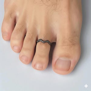 May include: A dark gray toe ring with a wavy design. The ring is worn on a toe, and the image is a close-up shot. The ring's design resembles a stylized wave.