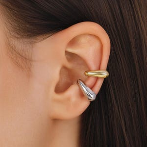 May include: A close-up of an ear adorned with two ear cuffs. One cuff is gold-toned and wraps around the upper ear, while the other is silver-toned and hangs from the ear's cartilage. The jewelry has a smooth, polished finish.