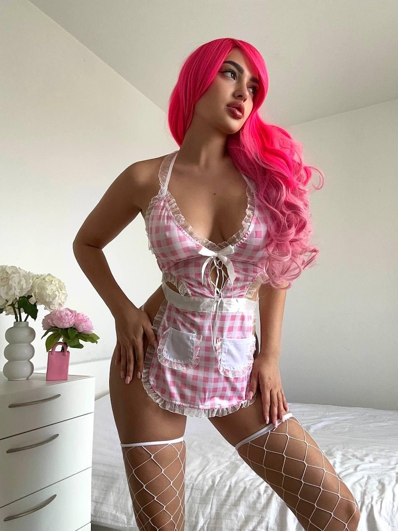 May include: A pink and white gingham lingerie set with a halter top and apron-style skirt. The set features white lace trim and a bow detail. White fishnet stockings complete the look. The model is wearing a pink wig.