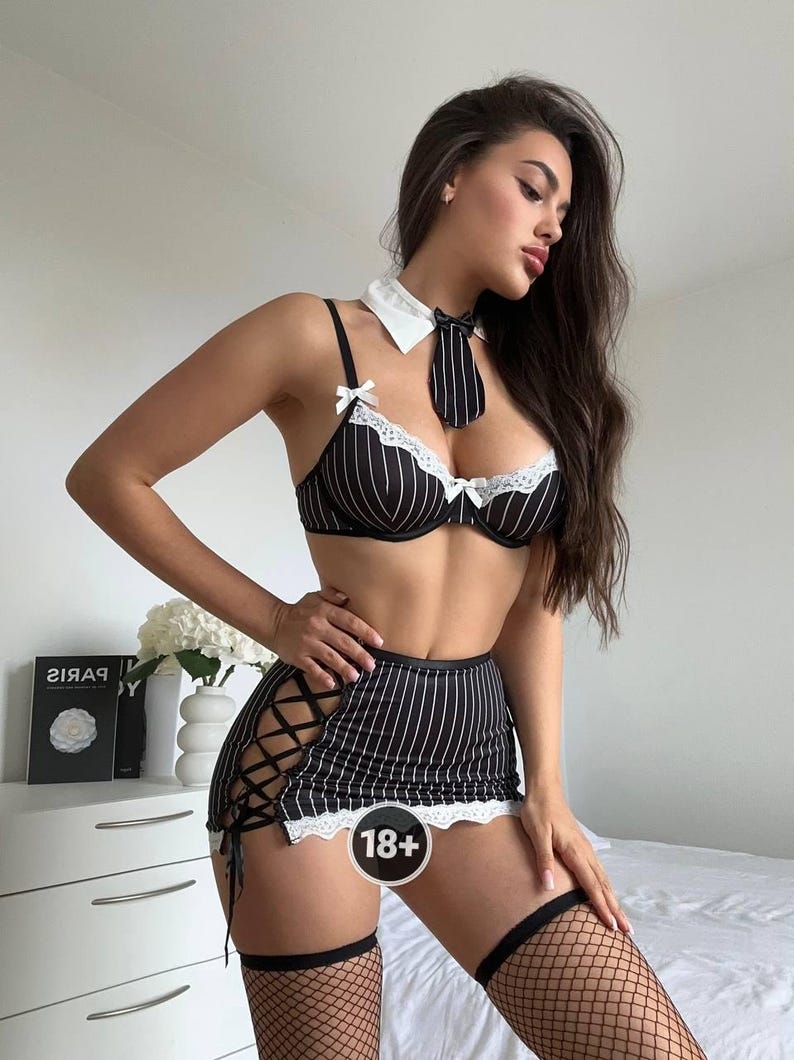 May include: Black and white pinstripe lingerie set with a matching tie and collar. The set includes a bra, skirt with lace-up detailing, and a mini skirt. The bra and skirt have white lace trim. The model is wearing black fishnet stockings.
