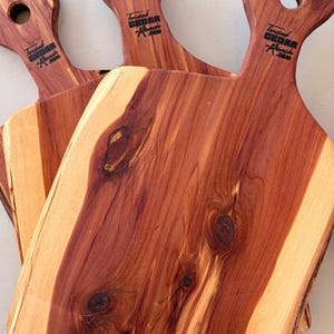 May include: Three wooden serving boards with handles, each featuring a circular hole for hanging. The boards display a natural grain pattern with reddish-brown centers and lighter cream-colored edges. Each handle is branded with Twisted Cedar Ranch.