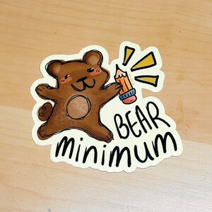 Puede incluir: Una pegatina con un oso de dibujos animados marrón sosteniendo un lápiz con rayos amarillos. El texto "BEAR minimum" está escrito en negro, con una fuente estilizada debajo del oso.