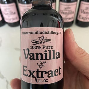 May include: A dark glass bottle of 100% Pure Vanilla Extract with a pink label. The label includes the text "Vanilla Extract" and the website address www.vanilladistillery.ca. The bottle is held in a hand.
