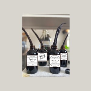 May include: Four dark glass bottles of vanilla extract with vanilla beans protruding. Each bottle has a white label with black text. The labels read "Handcrafted Vanilla Extract", "Homemade Vanilla Extract", and "Small Batch Vanilla Extract".