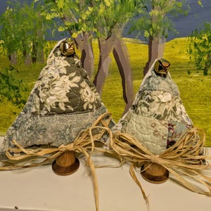 May include: Two handmade fabric Christmas tree decorations. The trees are made of quilted fabric with floral patterns in shades of green and cream. Each tree has a small gold bell at the top and is attached to a wooden spool base with a raffia bow.