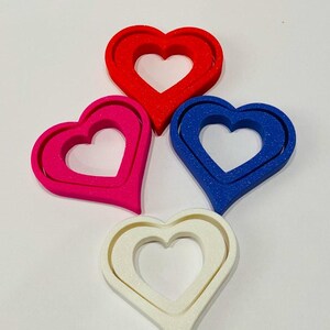 May include: Four heart-shaped cookie cutters, each with a double-layered design. The cutters are red, pink, blue, and white.