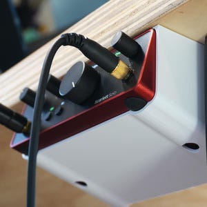 May include: A white and red audio interface, mounted under a wooden shelf. The interface has black knobs and inputs for cables. The text "Scarlett Solo" is printed on the device.