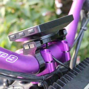 Puede incluir: Primer plano de un manillar de bicicleta con un dispositivo negro montado. El manillar es de color púrpura vibrante con el texto "WARP 9" en blanco. El dispositivo tiene varios botones y está montado con hardware negro.