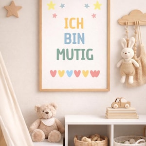 Könnte beinhalten: Ein gerahmter Druck mit dem deutschen Satz "ICH BIN MUTIG" in bunten Buchstaben, was "Ich bin mutig" bedeutet. Das Kunstwerk ist mit Sternen und Herzen verziert. Ein Teddybär, ein sternförmiges Kissen und andere Spielsachen sind in der Nähe.