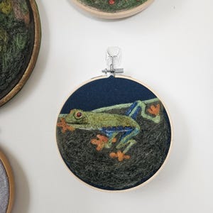 以下が含まれることがあります： 木製の刺繍枠の中に、オレンジ色の足と目を持つ緑と青のツリーフロッグを描いた手作りのフェルトアート作品。他の同様のアート作品が背景に見えます。