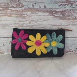 May include: A dark gray felt pouch with a zipper closure and a brown leather pull tab. The pouch is decorated with three large, colorful felt flowers in pink, yellow, and teal. The flowers have contrasting centers.