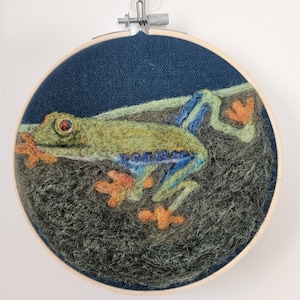 以下が含まれることがあります： 赤い目のアマガエルを描いた手作りの刺繍アート。 緑、青、オレンジの色合いで表現されたカエルは、濃い青色の布の背景に配置されています。 木製のフレームに収められた、壁掛けに適した作品です。