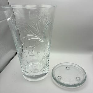 May include: A clear glass vase with etched floral designs and a matching round glass candle holder. The vase has a flared shape and is approximately 10 inches tall. The candle holder has three small holes.