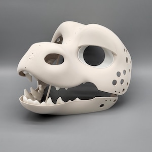 May include: A cream-coloured dinosaur mask with large eye openings and a wide open mouth revealing sharp teeth. The mask has several small holes on the sides and top, set against a grey background.