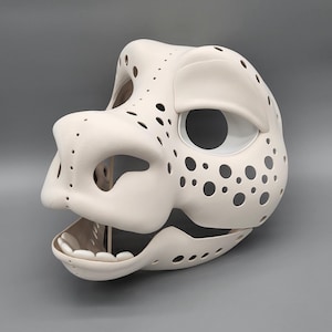 May include: A cream-coloured mask with an open mouth, large eye sockets, and numerous small, evenly spaced holes. The mask features a rounded snout and a smooth, matte finish. The design suggests a stylised animal or creature.
