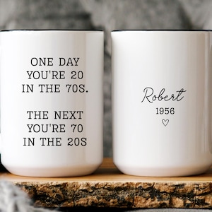 May include: Two white ceramic mugs with black handles and rims. The left mug features black text reading ONE DAY YOU'RE 20 IN THE 70S. THE NEXT YOU'RE 70 IN THE 20S. The right mug displays the name Robert, the year 1956, and a small heart symbol.
