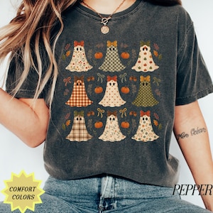May include: A charcoal gray short-sleeve t-shirt featuring a repeating grid pattern of nine ghosts. Each ghost has a unique pattern, including floral, plaid, checkered, and leaf designs, topped with a small bow. Small orange pumpkins are interspersed.