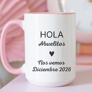 May include: A white ceramic mug with a light pink handle and interior rim. The front features black text that reads: HOLA Abuelitos, a small black heart icon, and the text: Nos vemos Diciembre 2026. The mug has a smooth, glossy finish.
