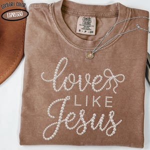 May include: Espresso-colored t-shirt with the phrase "loves like Jesus" in white pearl-like beads. A silver necklace with a small pendant rests on the shirt. The shirt has a crew neck and a Comfort Colors tag.