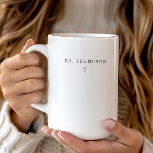 May include: A white ceramic mug with the text "DR. THOMPSON" and a heart symbol. The mug has a large handle and is held by a person wearing a cream-colored sweater.