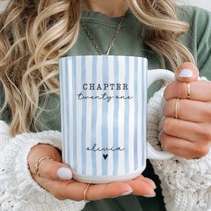 May include: A white ceramic mug featuring vertical light blue watercolor stripes. The text "CHAPTER twenty one" is printed in black, followed by the name "olivia" and a small black heart symbol centered on the front of the mug.