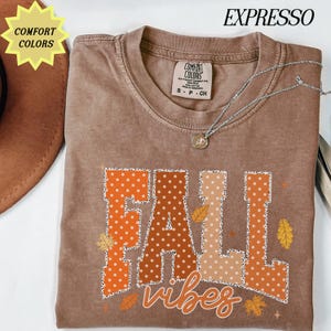 May include: A brown short-sleeve t-shirt with a crew neckline and a Comfort Colors brand label. The graphic features the text FALL in large block letters with white polka dots and the word vibes in script, surrounded by small autumn leaf icons.
