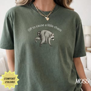 May include: A moss green short-sleeve t-shirt featuring a graphic of a raccoon walking on all fours. Above the raccoon, the white text reads, Off to cause a little chaos. The shirt has a crew neckline and a relaxed fit, showcasing a casual style.