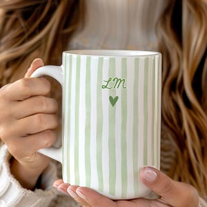 May include: A white ceramic mug featuring vertical light green stripes, the letters LM in a cursive green font, and a small green heart icon centered below the text. The mug has a curved handle on the left side and a smooth, cylindrical body.
