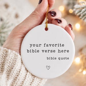 May include: A round white ceramic ornament featuring black printed text that reads your favorite bible verse here bible quote with a small hand-drawn heart symbol below the text. The ornament includes a gold string loop for hanging purposes.