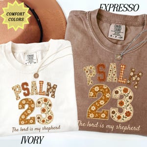 May include: Two Comfort Colors t-shirts in ivory and espresso, each featuring the text PSALM 23 and The lord is my shepherd. The letters and numbers are filled with floral and gingham patterns in brown, orange, and cream tones. A silver necklace rests on each.