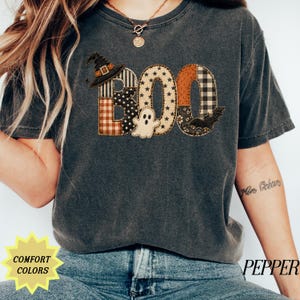 May include: A charcoal gray short-sleeve t-shirt featuring the word BOO in a patchwork style with various patterns including stars, plaid, and polka dots. A small witch hat sits on the B, a ghost is between the letters, and a bat is on the O.