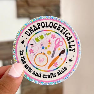 May include: A circular holographic sticker with a pink border and the text "UNAPOLOGETICALLY in the arts and crafts aisle". The center features illustrations of a green paint tube, a paint palette, pink scissors, a paintbrush, and an embroidery hoop.
