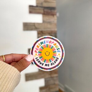 Pu&ograve; includere: Un adesivo circolare con un sole giallo centrale con un semplice volto sorridente nero, circondato da linee radianti multicolori. Il testo PERIMENOPAUSE LEAVE ME ALONE &egrave; stampato in lettere maiuscole nere attorno al disegno del sole.