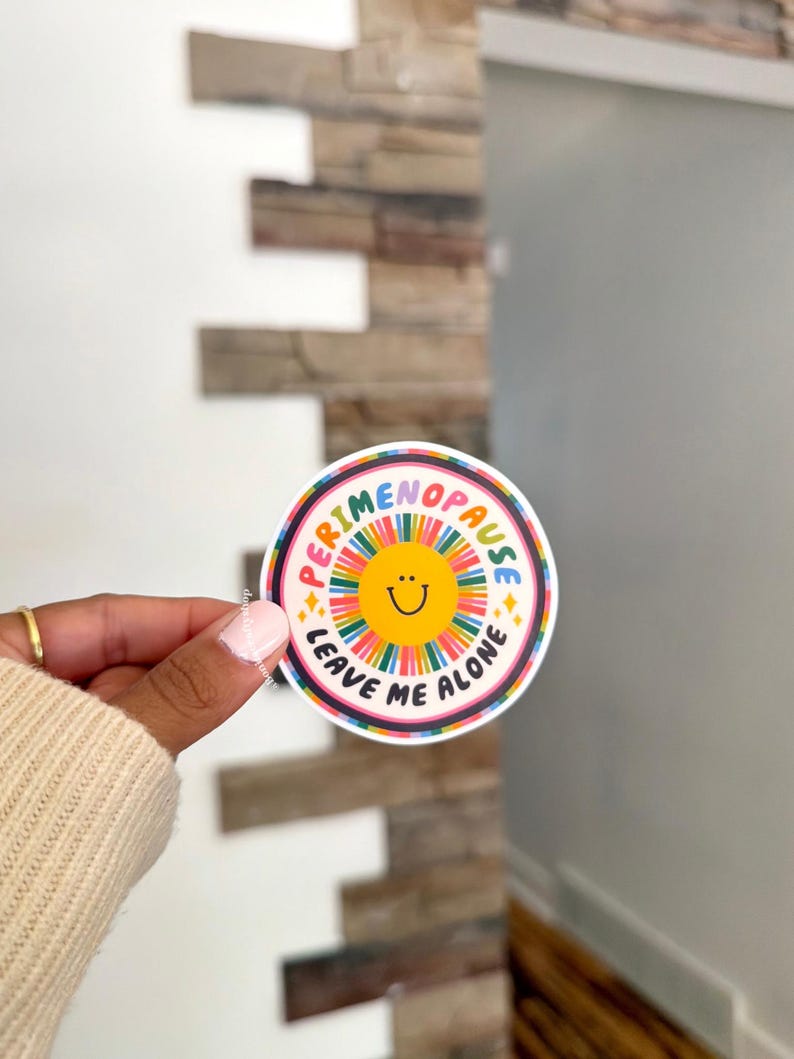 Pu&ograve; includere: Un adesivo circolare con un sole giallo centrale con un semplice volto sorridente nero, circondato da linee radianti multicolori. Il testo PERIMENOPAUSE LEAVE ME ALONE &egrave; stampato in lettere maiuscole nere attorno al disegno del sole.