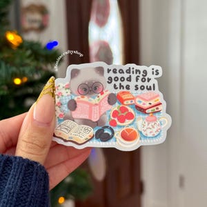 May include: A sticker featuring a cartoon cat wearing glasses and reading a book. The sticker includes the text "reading is good for the soul" and illustrations of books, strawberries, and tea.