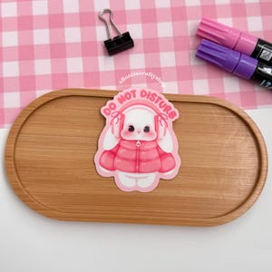 May include: A pink and white sticker featuring a cartoon bunny wearing a pink jacket and earmuffs, with the words "DO NOT DISTURB". The sticker is on a wooden tray, with pink and purple markers and a black binder clip.