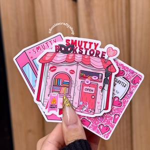 May include: A collection of pink stickers featuring a bookstore theme. One sticker depicts a pink building with the text "Smutty Bookstore" and a "Hot & Heavy" warning sign. Other stickers have hearts and the text "Chapter 14".