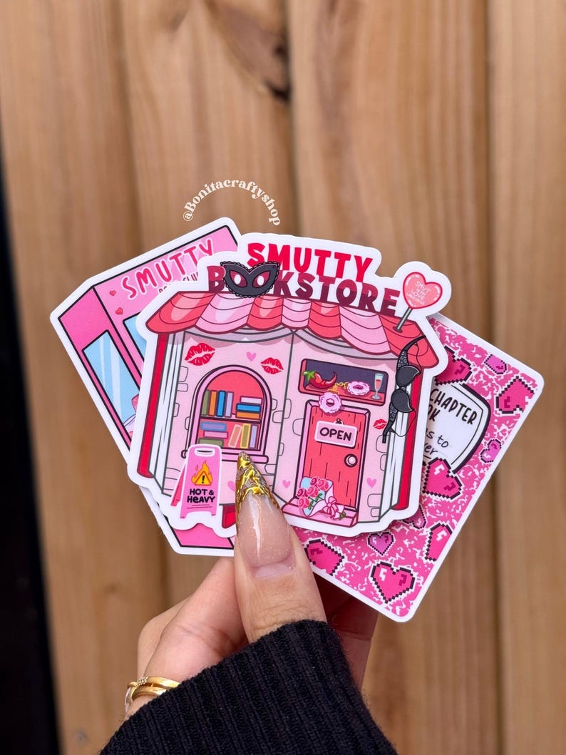 May include: A collection of pink stickers featuring a bookstore theme. One sticker depicts a pink building with the text "Smutty Bookstore" and a "Hot & Heavy" warning sign. Other stickers have hearts and the text "Chapter 14".