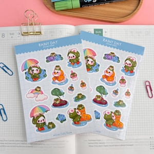 May include: Two sticker sheets with a "Rainy Day" theme. Each sheet features cute cartoon characters with umbrellas, rain boots, and flowers. The stickers are in various colors, including green, orange, and blue. Other stationery items are in the background.
