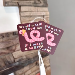 May include: Two square stickers with a maroon background and a pink worm illustration. The text on the sticker reads "would u still love me if I were a worm". The worm has a heart-shaped design and a yellow bandana.