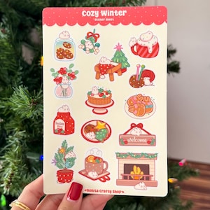 May include: A sticker sheet with a red border and the words "cozy Winter" at the top. The sheet features various winter-themed illustrations, including cookies, a Christmas tree, a mug of hot cocoa, and a fireplace. The bottom right corner has the text "Bonita Crafty Shop".