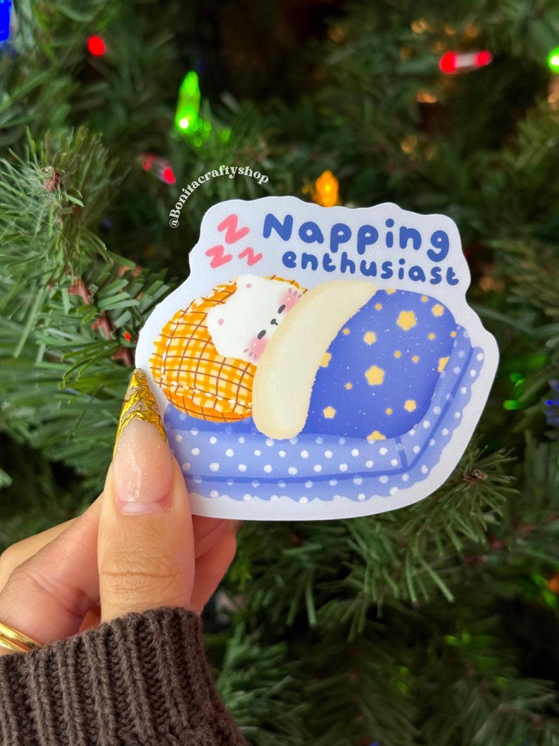 May include: A sticker with a cartoon illustration of a sleeping bear on a blue pillow with yellow stars. The sticker has the text "Napping enthusiast" in blue. The background is a Christmas tree.
